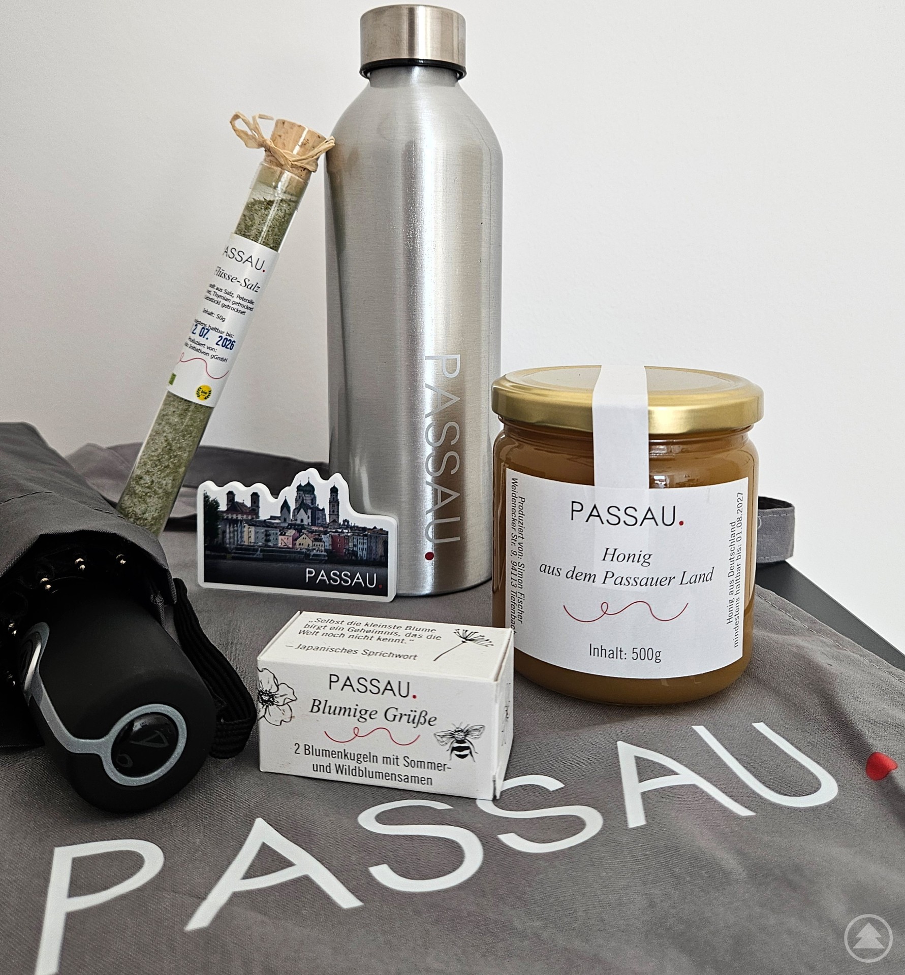 Die neuen nachhaltigen Souvenirs der Tourist-Information Passau. Auf dem Bild sind verschiedene Souvenirs der Stadt Passau zu sehen, darunter eine silberne Trinkflasche, ein Glas Honig, ein Taschenschirm, ein Magnet mit Stadtmotiv, ein Reagenzglas mit „3-Flüsse-Salz“ sowie eine kleine Box mit Blumenkugeln. Alle Produkte tragen das Branding „PASSAU.“.