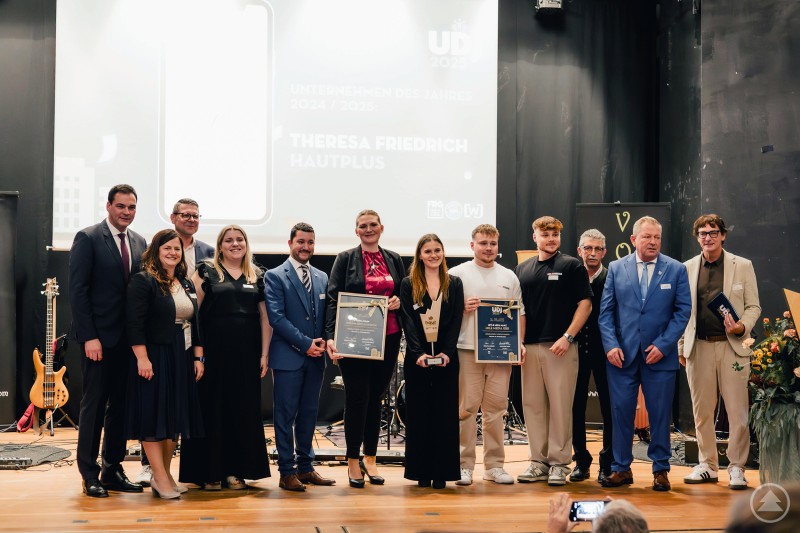 Freude über das Gewinnerpodium auf der Bühne im Kurhaus Freyung: Landrat Sebastian Gruber, Handwerkskammer-Vizepräsidentin Kathrin Zellner, IHK-Vizepräsident Jürgen Greipl, WJ-Vice President Anna Schraml, WJ-President Alexander Treml, Karina Haim (Leckere Köstlichkeiten), Theresa Friedrich (HautPlus), Mika und Levi Maaz (Mile Media), Johann Kapfer (3. Bürgermeister Waldkirchen), Alexander Pieringer (Bürgermeister Fürsteneck), Regionalmanager Stefan Schuster (von links) Eine Gruppe elegant gekleideter Personen steht auf einer Bühne vor einer dunklen Leinwand. Zwei der Personen in der Mitte halten gerahmte Auszeichnungen. Alle lächeln in Richtung Kamera, im Vordergrund ist ein Handy zu sehen, das die Szene fotografiert.