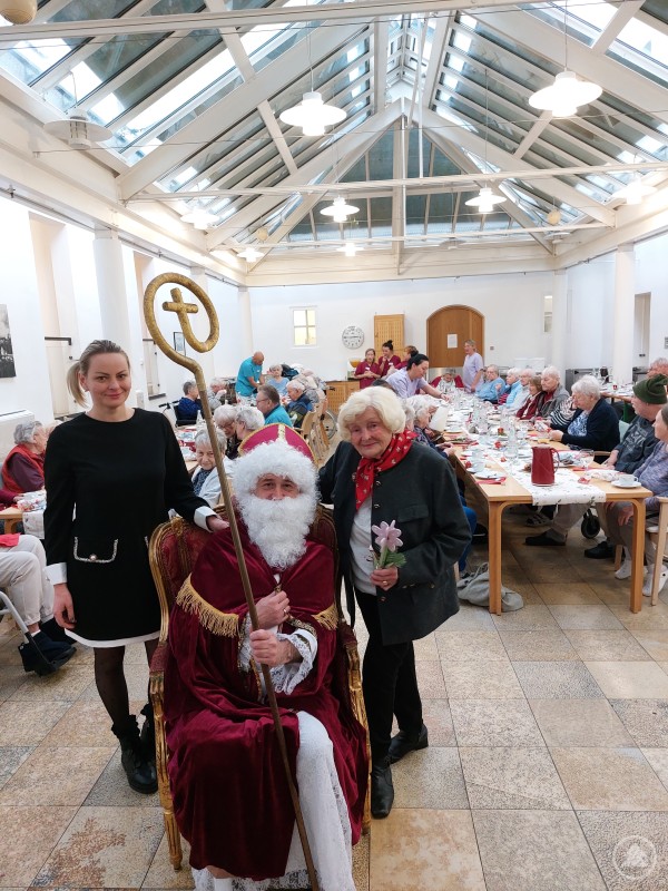 v.l. Die Pflegedienstleiterin des Seniorenstifts, Frau Rinata Deibele, gemeinsam mit dem Nikolaus und der ehrenamtlichen Verwaltungsrätin Hildegunde Brummer In einem lichtdurchfluteten Saal mit Glasdach sitzt eine große Gruppe Seniorinnen und Senioren an festlich gedeckten Tischen. Im Vordergrund stehen der Nikolaus mit Bischofsstab, eine ältere Dame mit Blume und die Pflegedienstleiterin. Alle wirken fröhlich und genießen die Feier.