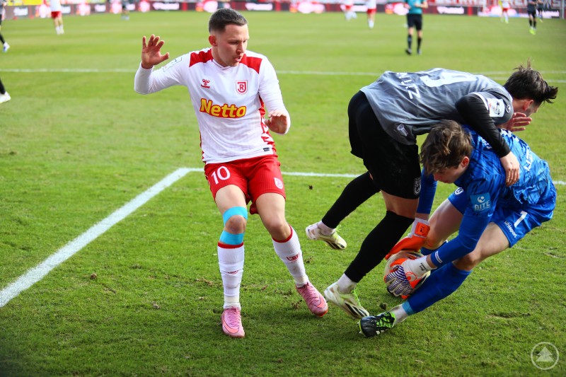 Ein Spieler des SSV Jahn Regensburg im wei&szlig; roten Trikot versucht im Strafraum an Torh&uuml;ter und Verteidiger des SSV Ulm vorbeizukommen, w&auml;hrend der Keeper den Ball sichert.