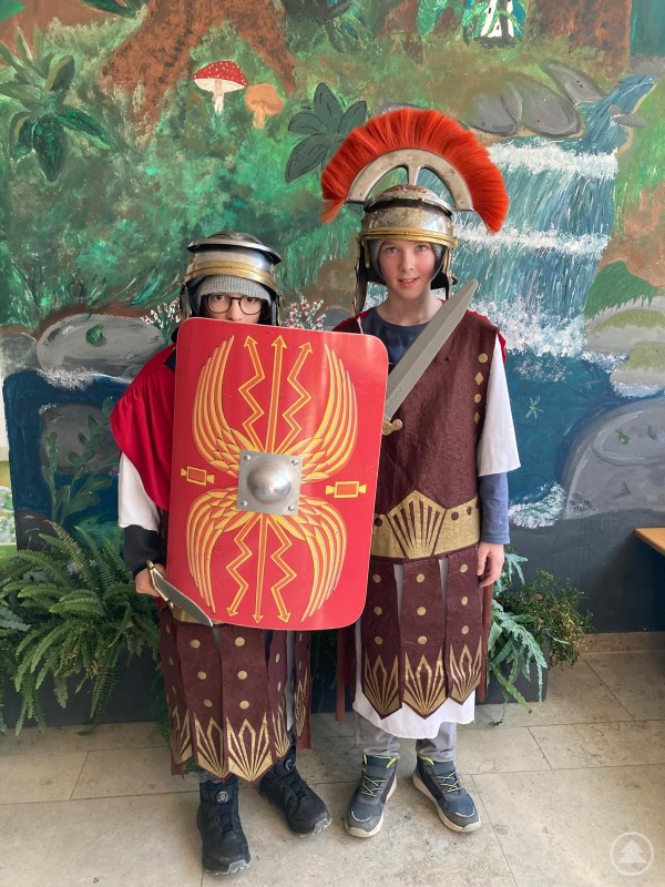Zwei Sch&uuml;ler stehen in r&ouml;mischer Verkleidung mit Helmen, rotem Schild und Schwert vor einer bemalten Kulisse.