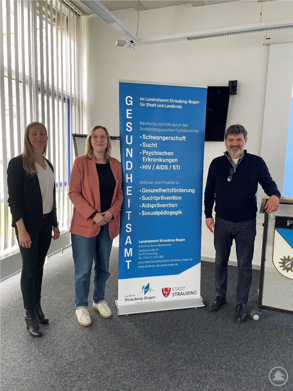 Alexandra Jehle vom Gesundheitsamt Straubing mit Vertretern des Polizeiverwaltungsamtes beim Vortrag zur Gesundheitsförderung Drei Personen stehen vor einem Informationsbanner des Gesundheitsamtes im Raum eines Behördengebäudes