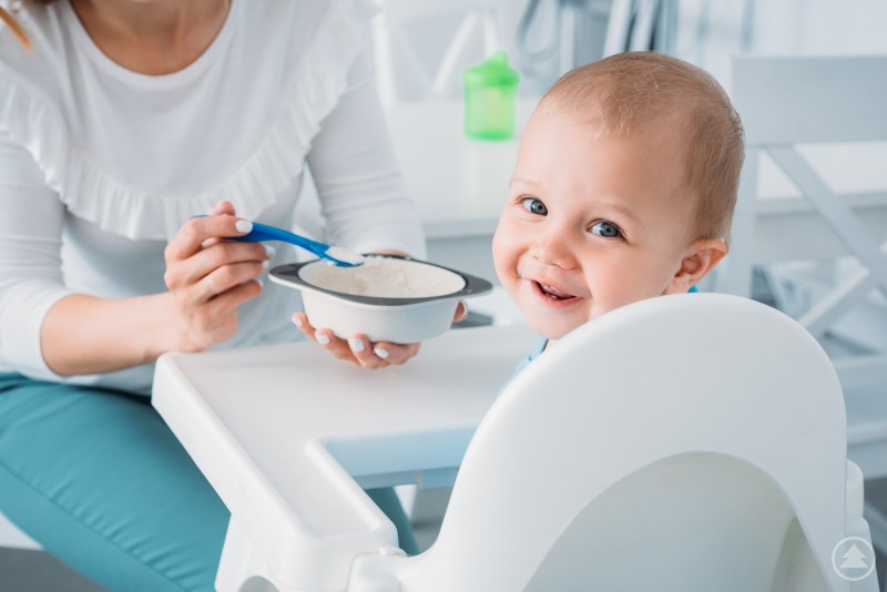 Gesundes Essen von Anfang an: Baby entdeckt die ersten Breimahlzeiten Ein lachendes Baby sitzt in einem weißen Hochstuhl und wird von einer erwachsenen Person mit Brei gefüttert. Die Szene vermittelt eine ruhige und freundliche Atmosphäre rund um gesunde Ernährung im Kleinkindalter.