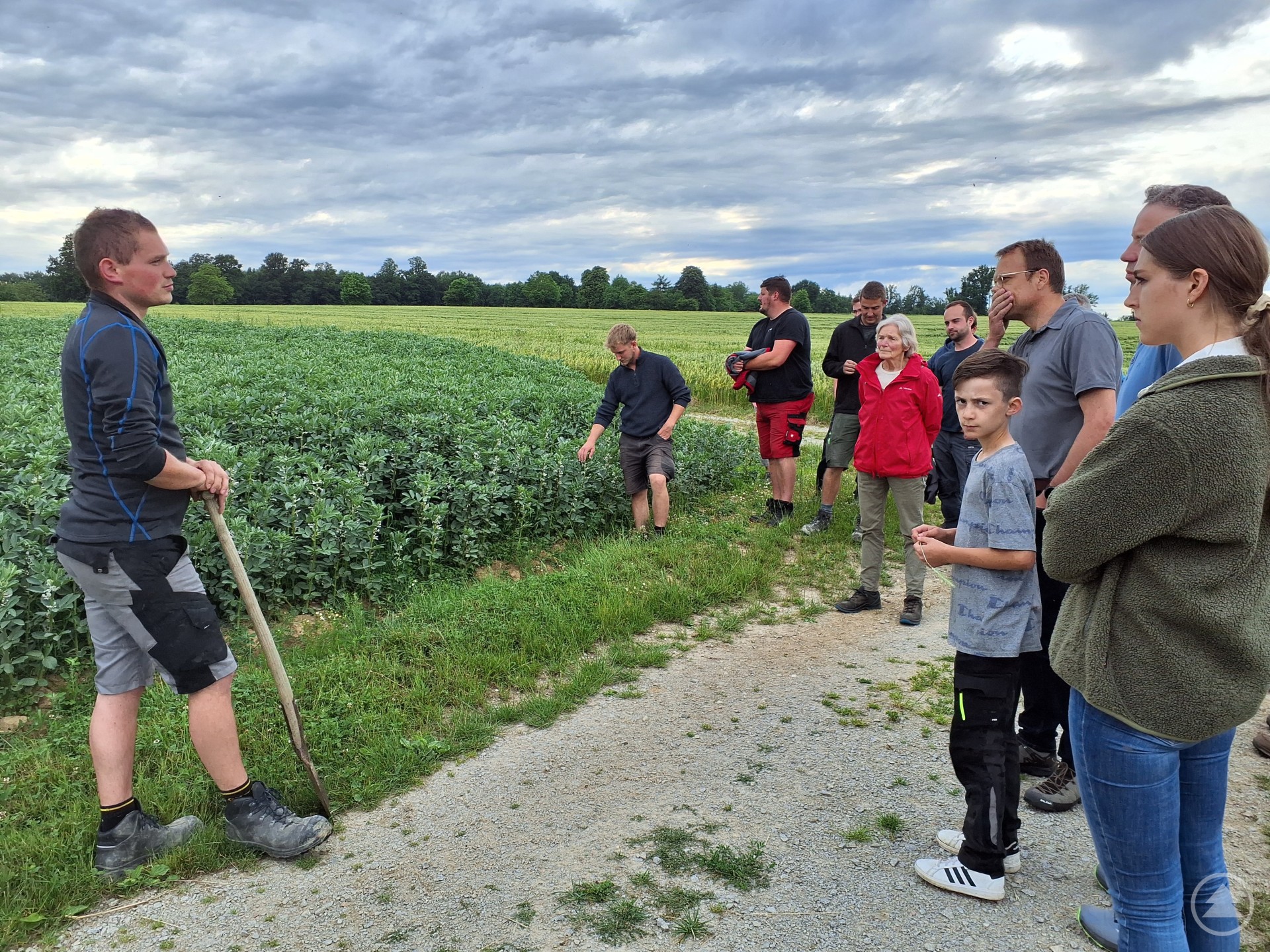 Ein Landwirt mit Hacke spricht vor einer Gruppe interessierter Personen am Feldrand. Im Hintergrund ist eine grüne Ackerfläche unter bewölktem Himmel zu sehen.