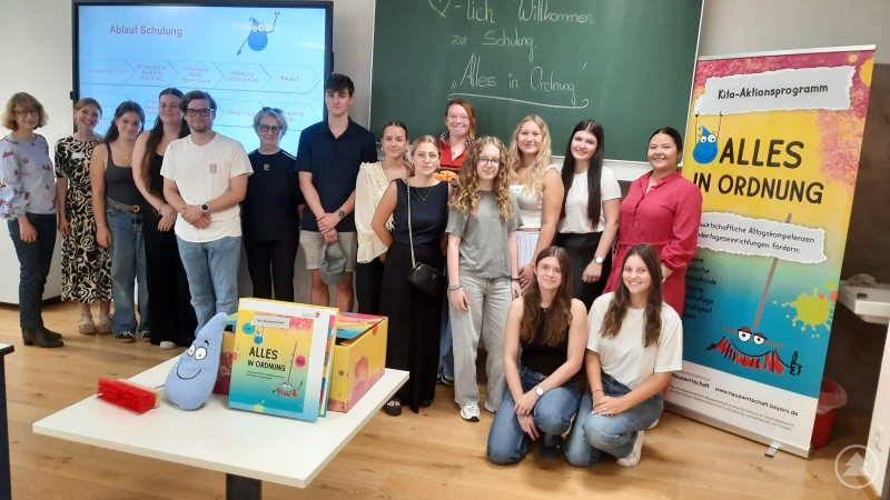 Dr. Tanja Steinberger (links) und Vanessa Handl (2. von links) freuten sich über das Interesse der künftigen Erzieherinnen und Erzieher Eine Gruppe junger Erwachsener steht gemeinsam mit zwei Lehrkräften vor einer Tafel und einem Roll-Up. Im Vordergrund sind Materialien und Maskottchen des Aktionsprogramms „Alles in Ordnung“ zu sehen.