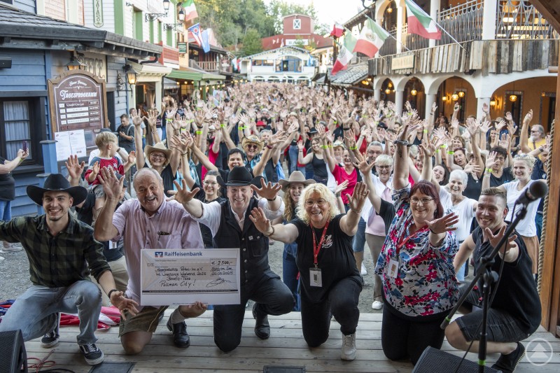 Strahlende Gesichter beim Benefiztanz: Josef Hofbauer von der Kinderkrebshilfe Rottal-Inn e.V. nimmt den Spendenscheck über 2.500 Euro dankend entgegen – gemeinsam mit hunderten Line Dancern in Pullman City. Eine große Menschenmenge jubelt auf der Main Street in Pullman City, vorne wird ein Scheck über 2.500 Euro an die Kinderkrebshilfe Rottal-Inn übergeben.