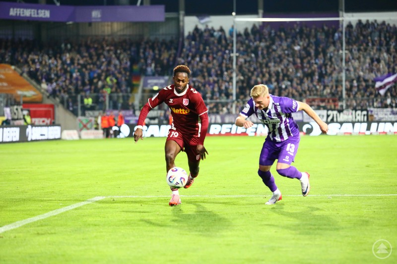 Schnelles Duell auf der Außenbahn beim Auswärtsspiel in Osnabrück. Ein Spieler des SSV Jahn Regensburg sprintet mit Ball am Fuß, während ihn ein Spieler des VfL Osnabrück eng verfolgt. Im Hintergrund sind die vollbesetzten Ränge zu sehen.