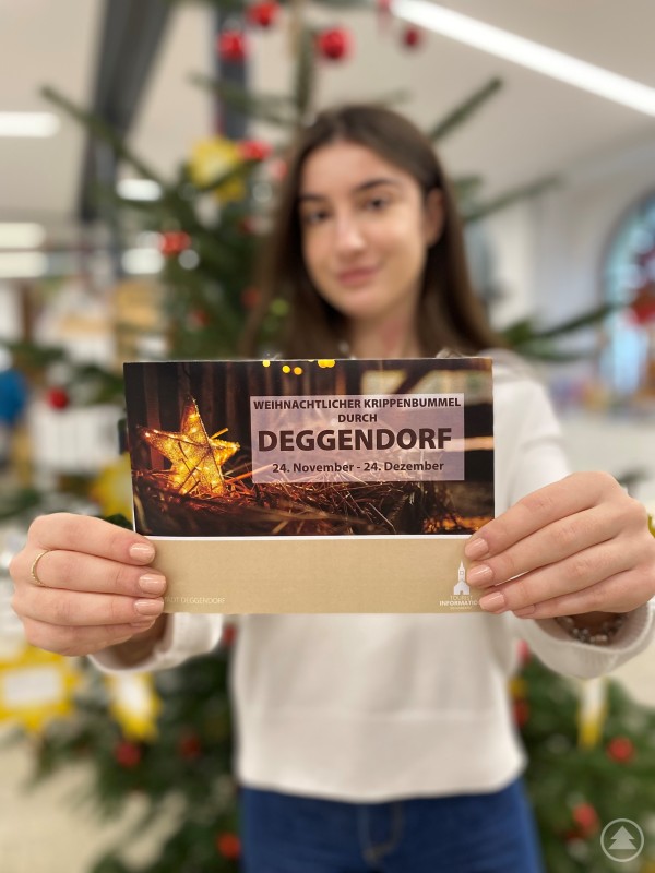 Praktikantin Erla freut sich, den Flyer ab sofort an Interessierte zu verteilen Eine junge Frau steht vor einem festlich geschmückten Weihnachtsbaum und hält einen Flyer zum „Weihnachtlichen Krippenbummel durch Deggendorf“ in die Kamera. Der Flyer zeigt einen beleuchteten Stern auf Stroh und die Veranstaltungsdaten.