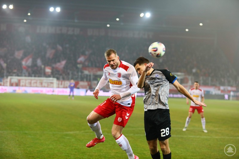 Zwei Spieler liefern sich ein Kopfballduell, im Hintergrund sind die voll besetzten Trib&uuml;nen des Jahnstadions zu sehen.
