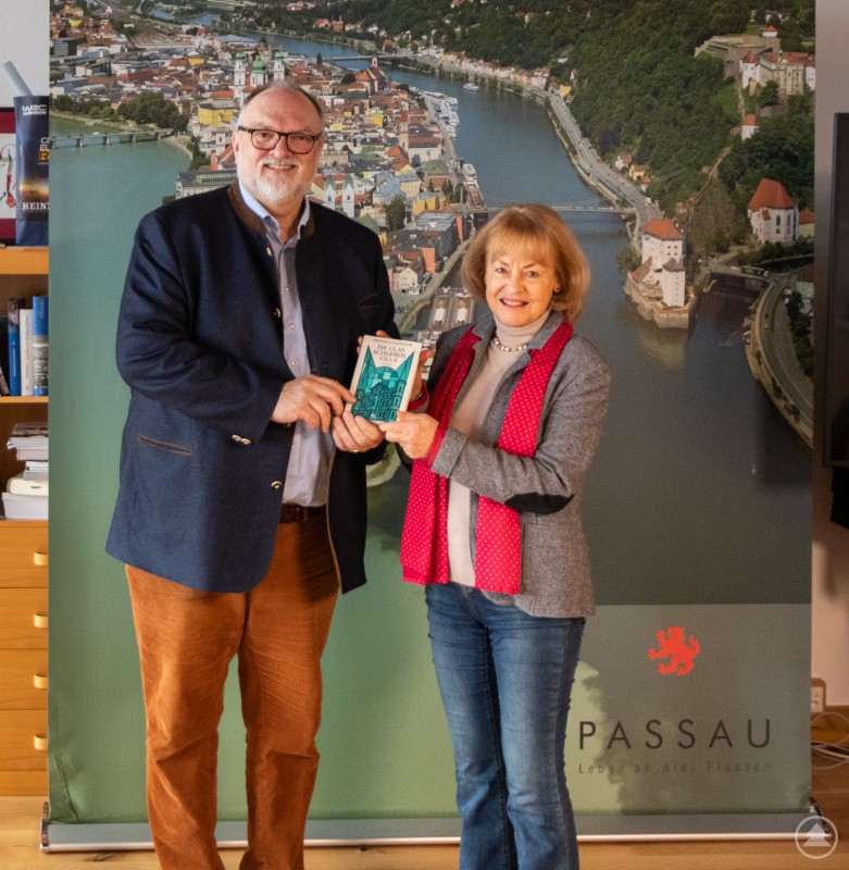 Autorin Monika Sadegor überreicht Oberbürgermeister Jürgen Dupper eines der ersten Exemplare ihres Romans „Die Glasscherbenvilla“ Eine Frau und ein Mann stehen vor einem großformatigen Luftbild von Passau. Die Frau überreicht dem Mann ein Buch und beide lächeln in die Kamera.