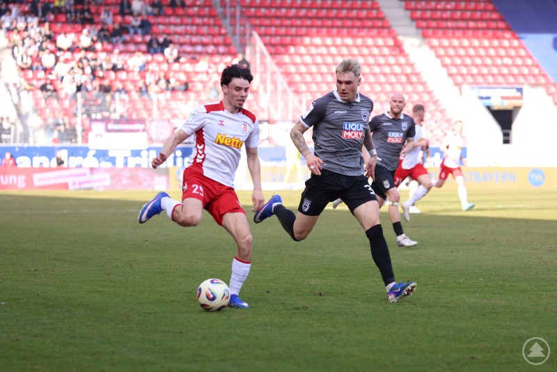 Intensives Duell zwischen dem SSV Jahn und dem SSV Ulm im Jahnstadion Regensburg. Ein Jahn Spieler im Sprintduell mit einem Ulmer Verteidiger, beide kämpfen um den Ball vor der Tribüne des Jahnstadions.