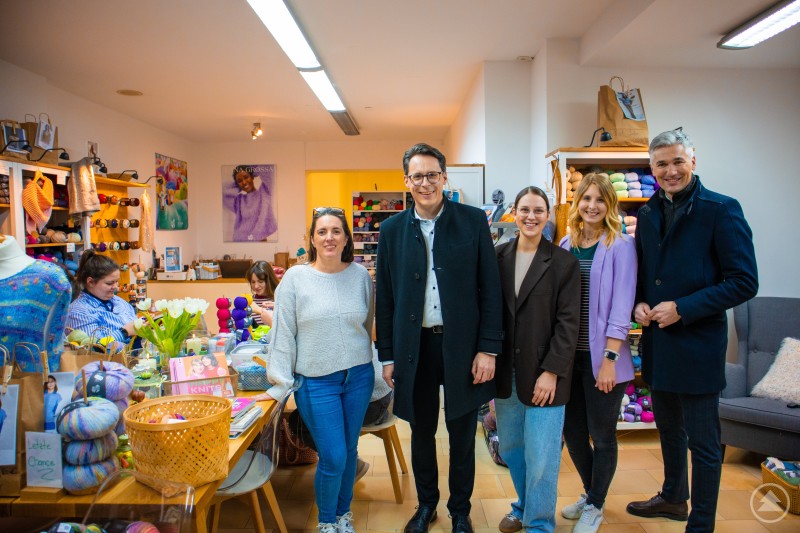 Ein Highlight des Abends: Das Wollgeschäft verwandelte sich in einen Treffpunkt für Strickbegeisterte, v.l. Stephanie Ries-Thomas, Geschäftsführung Wollkramerin; Oberbürgermeister Dr. Christian Moser; Franziska Krauth, Geschäftsführung Modehaus Krauth; Lisa Peter und Andreas Höhn vom Stadtmarketing Verein Mehrere Personen stehen in einem Woll- und Handarbeitsgeschäft zwischen Regalen voller bunter Wolle und Strickzubehör. Im Hintergrund sitzen Besucherinnen an einem Tisch und stricken gemeinsam. Das Geschäft ist hell beleuchtet und zeigt eine gemütliche Atmosphäre während einer Abendveranstaltung.
