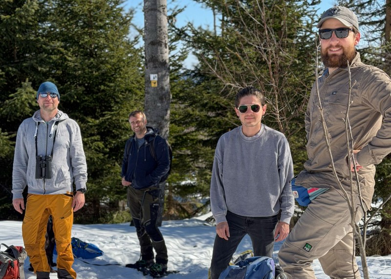 Für die erste Podcast-Folge ging es im Februar zusammen mit Forschern und viel Ausrüstung auf einen Auerhahnbalzplatz bei Finsterau. Vier Personen stehen mit Rucksäcken und Ausrüstung im Schnee auf einer Holzplattform im Wald. Im Hintergrund sind Nadelbäume zu sehen.