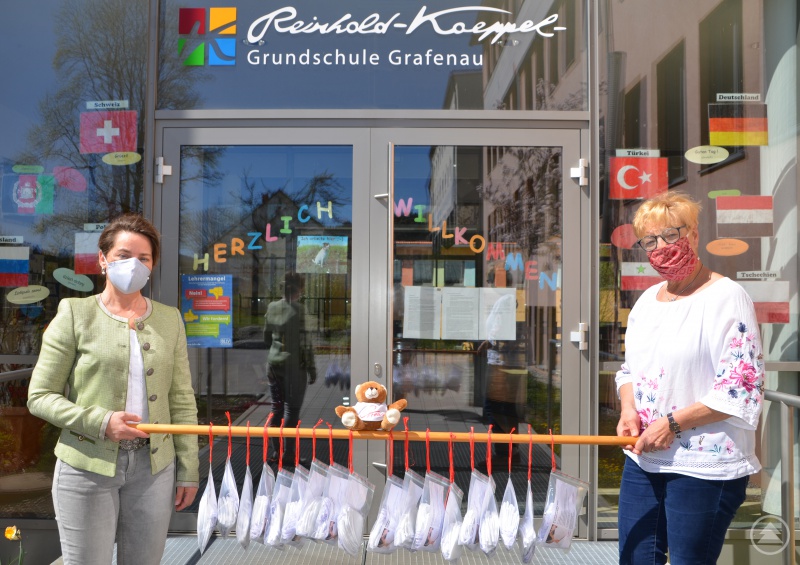Ungewöhnliche Zeiten erfordern eine ungewöhnliche Spendenübergabe: Rektorin Christl Weiß (re.) und Unternehmerin Maria Freund hielten bei der Spendenübergabe den Sicherheitsabstand geschützt durch Masken und durch eine zwei Meter lange Holzstange ein. Auf der Stange hingen mit roten Bändern die 175 gespendeten Masken mit einem Freund-Teddybären für die Grundschule Grafenau.