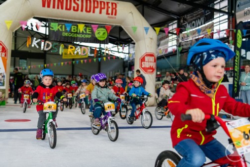 Start frei für die jüngsten Rennfahrer: Mit viel Begeisterung und bunten Helmen gingen die Kinder beim KiDS RADL RACE im Eisstadion Deggendorf an den Start – wetterbedingt wurde die Veranstaltung kurzerhand nach drinnen verlegt. Eine große Gruppe von Kleinkindern mit bunten Helmen fährt auf Laufrädern und Fahrrädern durch das Eisstadion Deggendorf. Sie starten unter einem aufblasbaren Bogen mit der Aufschrift „Windstopper“.