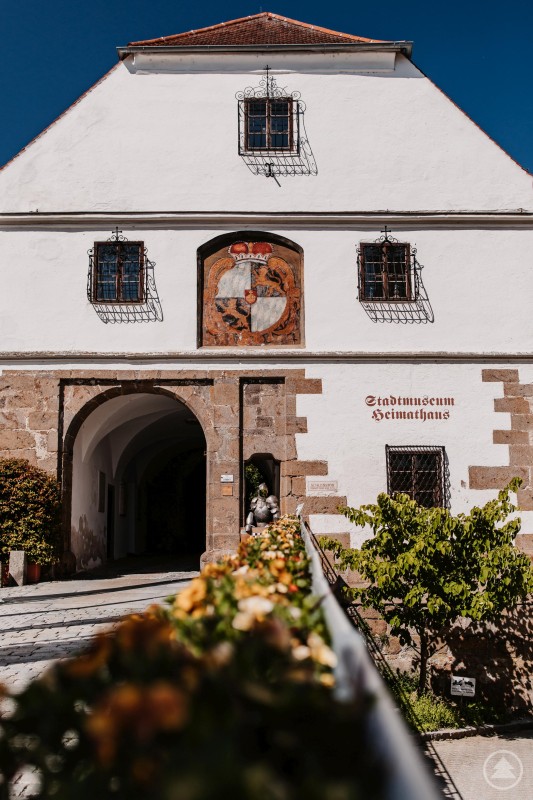 Das Stadtmuseum Schärding lädt im Juli zum kreativen Ferienprogramm mit dem Workshop „Bänderknüpfen mit Gerti“. Historisches Gebäude mit weißer Fassade, Rundbogenportal und Wappen über dem Eingang. Der Schriftzug „Stadtmuseum Heimathaus“ ist an der Wand angebracht. Im Vordergrund ein Geländer mit Blumenschmuck.