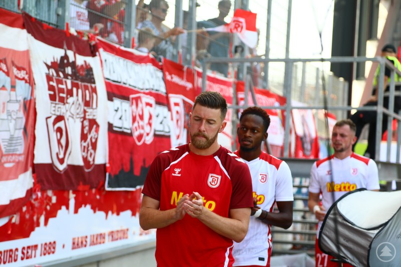 Spieler des SSV Jahn Regensburg laufen vor einer Tribüne mit rot-weißen Fanbannern und Fahnen ins Stadion ein.