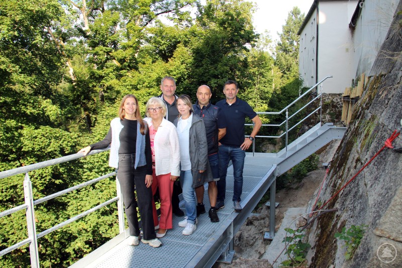 Auf dem neuen Wartungssteg an der südwestlichen Schlossmauer von Schloss Wolfstein (von links): Isabella Pauli, Helga Weinberger, Josef Burghard, Susanne Bauer, Bernhard Brandl und Reinhard Tolksdorf. Sechs Personen stehen auf einem neu errichteten Metallsteg mit Geländer vor der Schlossmauer von Schloss Wolfstein. Im Hintergrund sind Bäume und Teile der Anlage sichtbar.