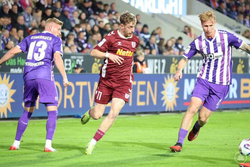 Ein Spieler des SSV Jahn Regensburg im Dribbling, bedrängt von zwei Osnabrücker Gegenspielern im Strafraumbereich.