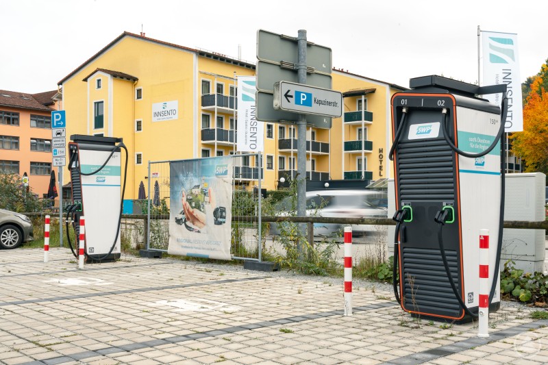 Der Park+Ride-Parkplatz in der Kapuzinerstraße ist mit zwei Schnellladern der Stadtwerke Passau einer der leistungsstärksten Ladepunkte im Stadtgebiet. Zwei moderne Schnellladestationen der Stadtwerke Passau stehen auf einem gepflasterten Parkplatz in der Kapuzinerstraße. Im Hintergrund sind ein gelbes Gebäude mit dem Schriftzug „INNSENTO“ sowie Hinweisschilder und Banner der Stadtwerke zu sehen.