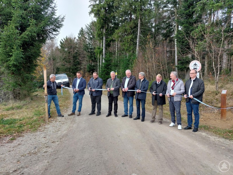 Auf die Loipe fertig los: die Bürgermeister eröffnen mit dem symbolischen Bandzerschneiden. (v.l.) Martin Eiberweiser (Ski & Bike), Bürgermeister Michael Reitberger (Lalling), Bürgermeister Robert Bauer (Schaufling), Bürgermeister Walter Nirschl (Bischofsmais), Bürgermeister Anton Stettmer (Grafling), Landrat Bernd Sibler (Deggendorf), zweiter Bürgermeister Günther Pammer (Deggendorf), stellvertretender Landrat Helmut Plenk (Regen) und MdL Martin Behringer (FWG). Eine Gruppe von neun Personen steht auf einer Waldstraße und durchschneidet gemeinsam ein Band zur feierlichen Eröffnung. Hinter ihnen sind Bäume und ein geparktes Auto zu sehen.