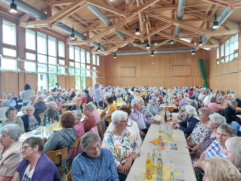 Gro&szlig;er Saal mit mehreren langen Tischen, an denen zahlreiche Seniorinnen und Senioren dicht beieinandersitzen. Die Josef-Eder-Halle ist gut gef&uuml;llt, es herrscht eine gesellige Atmosph&auml;re mit Gespr&auml;chen und Getr&auml;nken auf den Tischen.