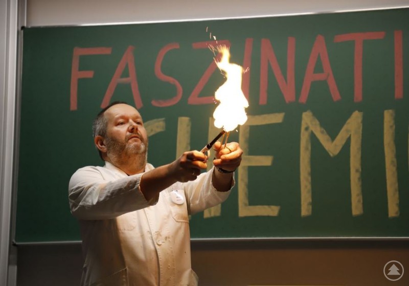 Ein Lehrer f&uuml;hrt im Klassenzimmer ein Experiment mit einer gro&szlig;en Flamme vor, im Hintergrund eine Tafel mit der Aufschrift Faszination Chemie.