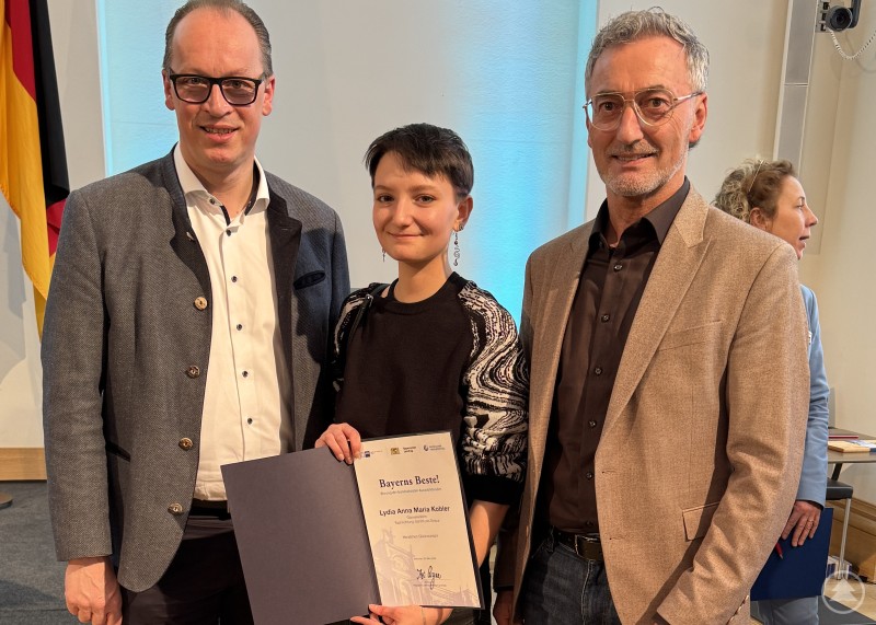 Lydia Kobler wurde im Maximilianeum für ihre herausragende Leistung als Glasveredlerin ausgezeichnet und erhielt persönliche Glückwünsche von MdL Martin Behringer. Drei Personen stehen bei einer Ehrung im Maximilianeum. In der Mitte hält eine junge Frau eine Urkunde, flankiert von zwei Männern in Anzügen.