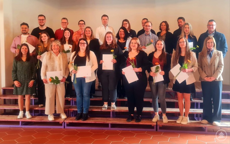 Hier sind die Absolventen zusammen mit Vertretern der Einrichtungen Gruppenfoto von Absolventinnen und Absolventen eines Pflege-Weiterbildungskurses zusammen mit Dozenten und Vertretern von Einrichtungen auf einer Bühne, viele halten Urkunden und Blumen in der Hand.