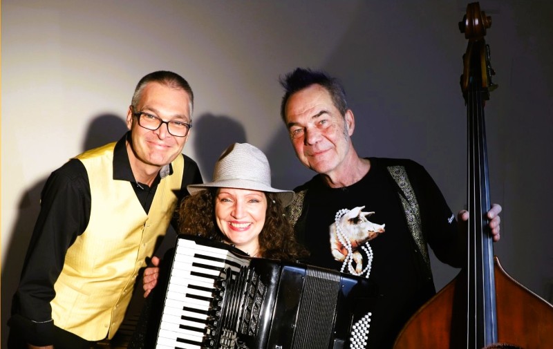 Drei Musiker stehen gemeinsam vor neutralem Hintergrund. Eine Frau mit Hut spielt Akkordeon, daneben stehen zwei Männer, einer mit Kontrabass. Alle lächeln in die Kamera.