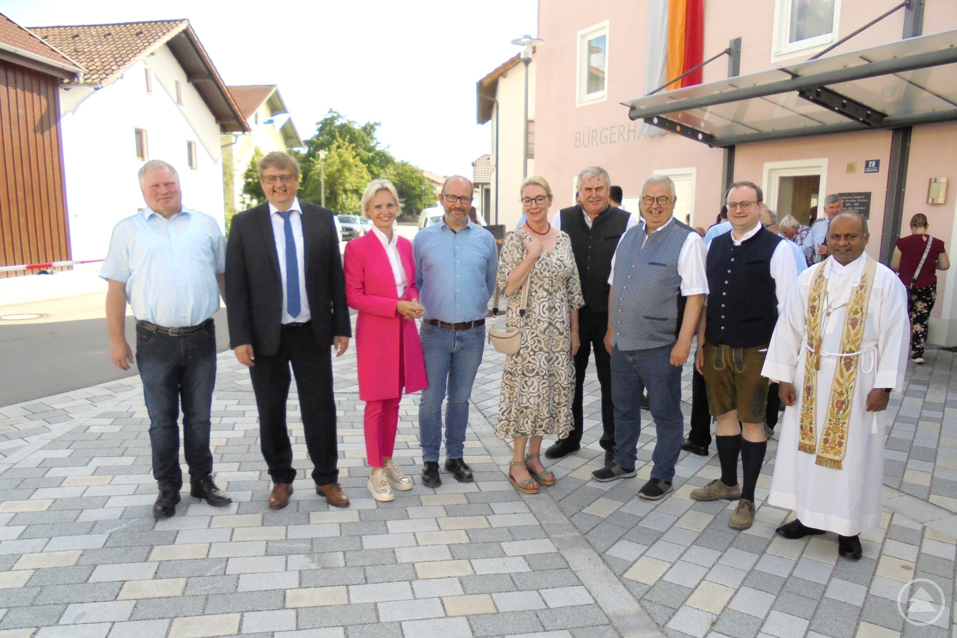 Über die sehr gelungene Umgestaltung der Ortsmitte von Uttigkofen freuen sich (v.l.): Josef Lechner, Hans-Peter Schmucker, Cornelia Wasner-Sommer, Florian Riesinger, Sabine Schmalhofer, Karl Haslinger, Harald Mayrhofer, MdL Stefan Meyer und Kaplan Pradeesh Antony Chullickal. Gruppe von zehn Personen, darunter Männer und Frauen in festlicher Kleidung, stehen lächelnd auf einem neu gestalteten gepflasterten Dorfplatz vor einem Gebäude mit Glasvordach und Deutschlandfahne.