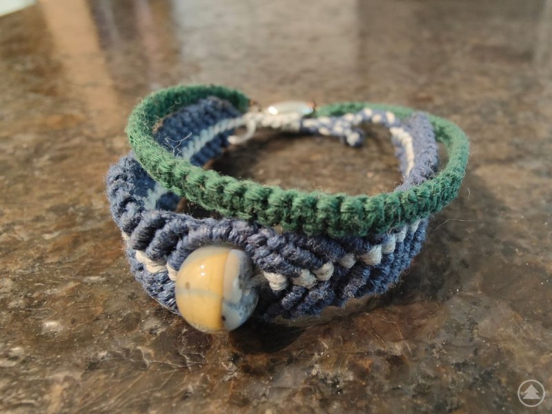 Handgefertigtes, geknüpftes Armband in Blau-, Grün- und Beigetönen mit einer dekorativen Perle, liegend auf einer dunklen, glänzenden Oberfläche.