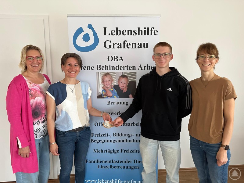 Alexander Fürst übergibt im Namen der Deutsch-Französisch-Klasse des LLG eine Spende an die OBA der Lebenshilfe Grafenau. Mit auf dem Foto: Naglmeier Annette, Daniela Springer und Veronika Fürst (v.l.). Vier Personen stehen lächelnd vor einem Roll-Up der Lebenshilfe Grafenau. Von links nach rechts: Annette Naglmeier, Daniela Springer, Alexander Fürst und Veronika Fürst. Alexander Fürst überreicht einen Geldschein als Spende.