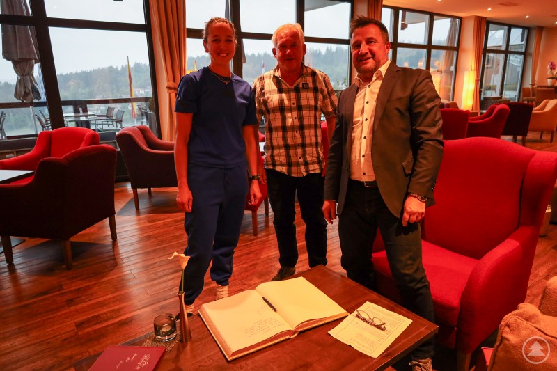 Eintrag ins Goldene Buch: Franziska Preuß, Herbert Unnasch und Bürgermeister Karl-Heinz Eppinger bei der Biathlon-DM in Zwiesel. Drei Personen stehen in einem Hotel-Loungebereich neben einem aufgeschlagenen Goldenen Buch. Eine Frau in Sportkleidung, ein Mann in kariertem Hemd und ein weiterer im Anzug lächeln in die Kamera.