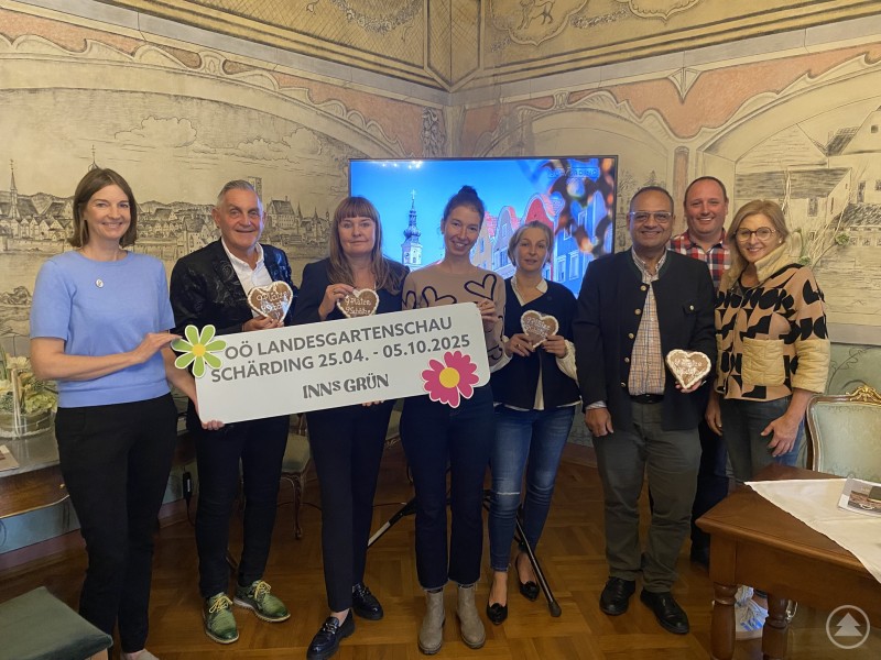 Andrea Berghammer, Bürgermeister Günter Streicher, Bettina Berndorfer, Nadia Musilek, Sandra Hofbauer, Maher Azer, Markus Furtner und Rosemarie Kaufmann (v.l.n.r.) Acht Personen stehen gemeinsam in einem historischen Raum mit Wandmalereien. Sie halten ein Schild zur Oberösterreichischen Landesgartenschau 2025 in Schärding sowie Lebkuchenherzen mit der Aufschrift „9 Plätze 9 Schätze“.