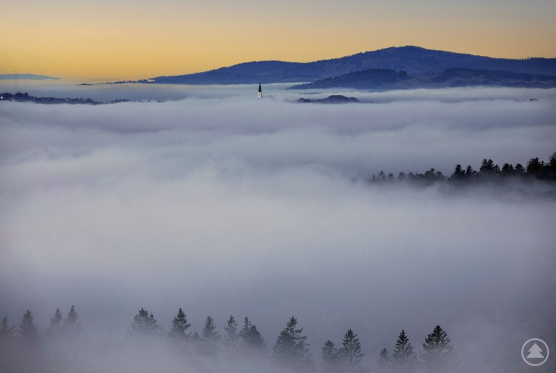 Das Foto zeigt einen Fotoblick gegen die Ausläufer des Rachel, davor lässt sich der Kirchturm von Kreuzberg erkennen, wie er aus dem Nebelmeer sticht. Auf dem Bild ist eine weite Landschaft zu sehen, die fast vollständig von dichtem Nebel bedeckt ist. Aus dem Nebel ragen die Spitzen von Nadelbäumen hervor, im Hintergrund sind sanfte Hügel oder Berge erkennbar. In der Ferne sticht ein Kirchturm aus dem Nebel heraus, darüber ein ruhiger Himmel in warmen Farben der Morgen- oder Abenddämmerung.