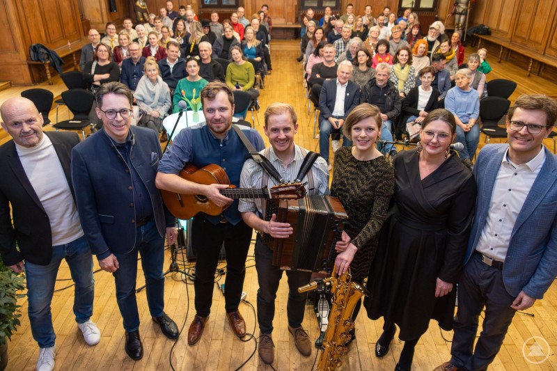 v.l.n.r. vorne: Konrad Rankl, Oberbürgermeister Dr. Christian Moser, die Großstadt Boazn mit Benedikt Landenhammer, Matthias Pürner und Birgit Tomys, Karin Loibl und Paul Linsmaier mit den geladenen Gästen im Hintergrund. Eine große Gruppe von Menschen sitzt in einem festlichen Saal. Im Vordergrund stehen mehrere Personen – darunter Musiker mit Gitarre, Akkordeon und Saxophon – gemeinsam für ein Gruppenfoto. Im Hintergrund sitzen viele Gäste und blicken zur Kamera.