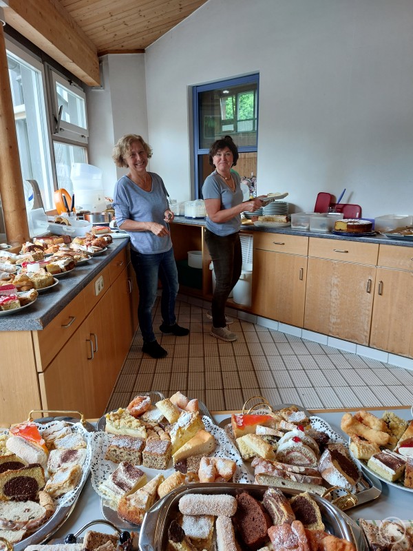 Die Seniorenbeauftragten im Ilzer Land kümmern sich auch in Hutthurm wieder selbst um das üppige Kuchenbuffet – da bleibt kein Wunsch offen. Zwei Frauen stehen hinter einem großen Kuchenbuffet in einer Halle. Vor ihnen sind zahlreiche selbstgebackene Kuchen und Torten auf Tabletts angerichtet, die für die Gäste vorbereitet werden.
