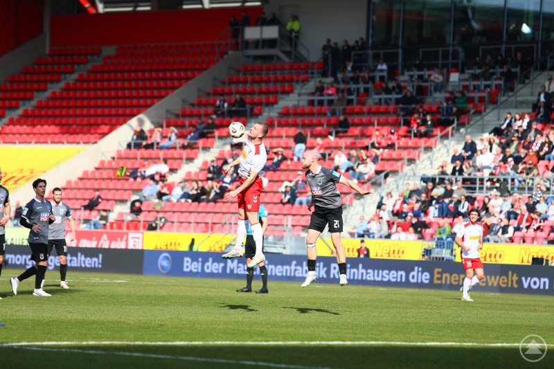 Zwei Spieler steigen im Mittelfeld zum Kopfball hoch, im Hintergrund die rot gef&uuml;llten R&auml;nge des Jahnstadions.