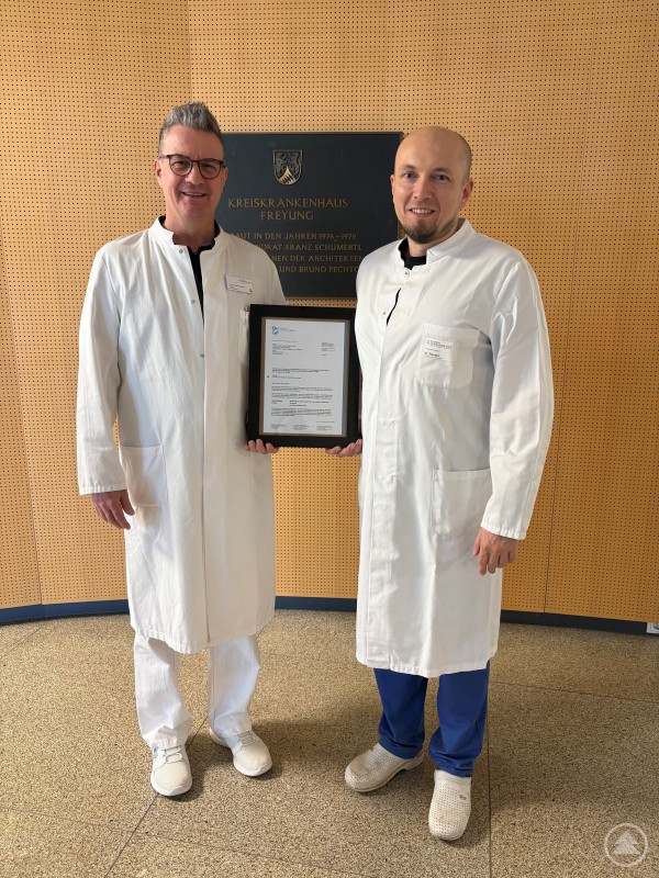 Die beiden weiterbildungsbefugten Ärzte Dr. Robert Ravasz und Dr. Michael Breitbach freuen sich über die erhaltene Weiterbildungsbefugnis Zwei Ärzte in weißen Kitteln stehen im Eingangsbereich des Kreiskrankenhauses Freyung vor einer Gedenktafel. Sie halten gemeinsam eine gerahmte Urkunde in den Händen und lächeln in die Kamera.