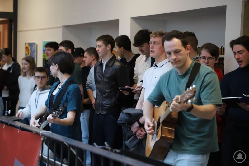 Eine Sch&uuml;lergruppe steht auf einem Flur und musiziert gemeinsam mit Gitarren, w&auml;hrend weitere Sch&uuml;ler im Hintergrund zuh&ouml;ren und mitsingen.